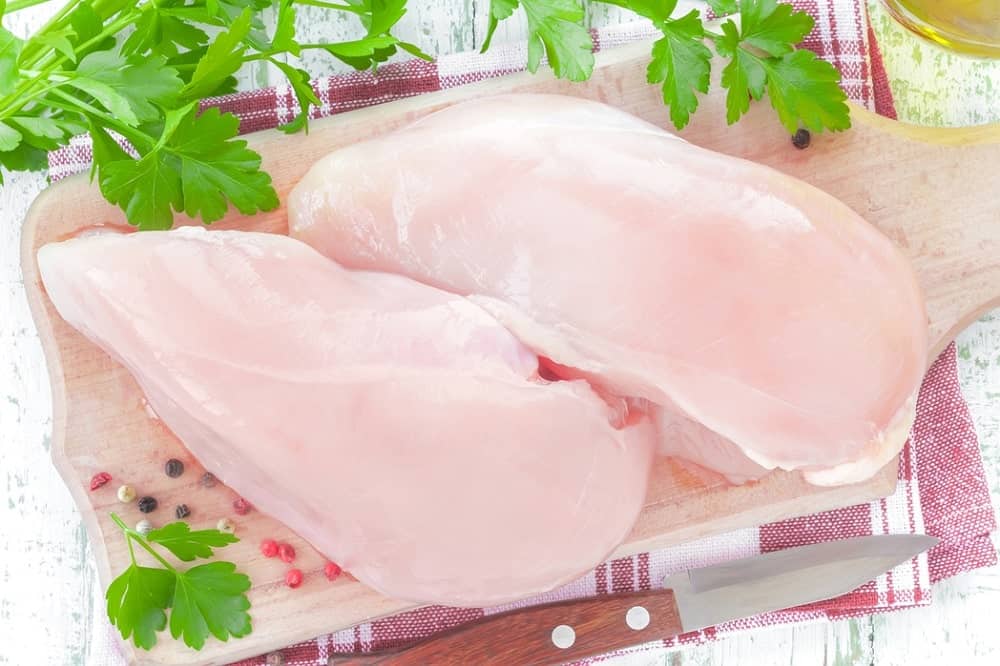 Raw chicken breasts, low in fat and calories, placed on a wooden chopping board with some herbs and spices on the side.